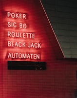 Red neon letters are mounted on a dark tiled wall, displaying the words Poker, Sic Bo, Roulette, Black Jack, and Automaten.