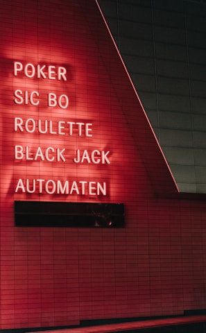 Red neon letters are mounted on a dark tiled wall, displaying the words Poker, Sic Bo, Roulette, Black Jack, and Automaten.
