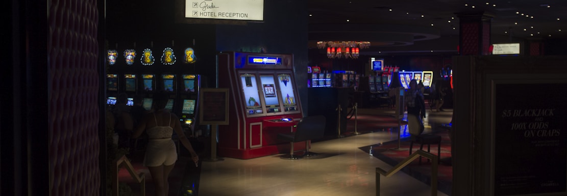 A dimly lit casino with several rows of slot machines, some displaying bright graphics and lights. There are a few people scattered throughout the area, engaging with the machines. The floor is reflective and polished, adding to the ambiance, while the ceiling is dotted with numerous small lights.