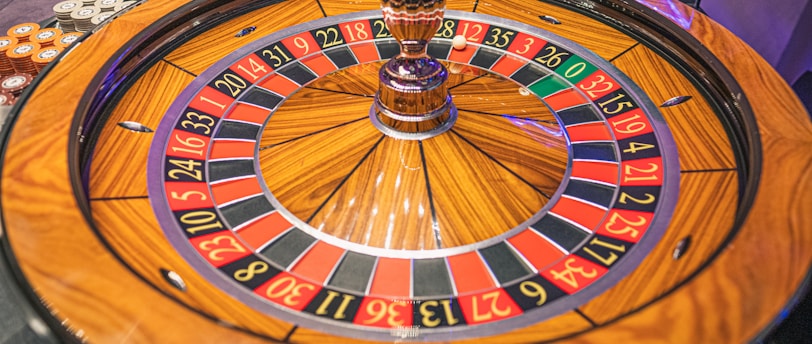 A roulette wheel with alternating red and black numbered pockets on a wooden platform. The wheel contains a small white ball resting in one of the pockets, indicating gameplay. Surrounding the wheel are stacks of colorful poker chips, suggesting a casino environment.