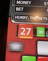 A digital display screen showing a casino betting interface, with text prompting to hurry bets and a highlighted number 27 in red. There are options for betting amounts and a 'share bets' button.