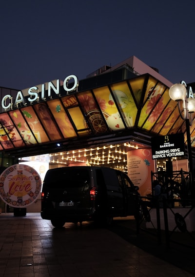 A building with a brightly illuminated entrance is seen at night. The sign above reads 'Casino' in bright letters. The facade features colorful graphics depicting various indulgent themes. Several vehicles are parked near the entrance, and people are visible in the vicinity. Tall lamp posts with spherical lights are present, casting a warm glow.