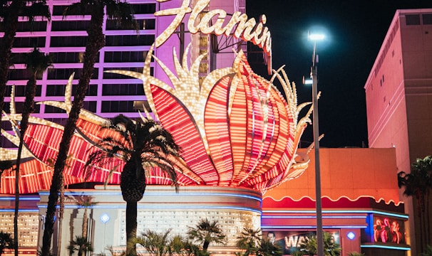 A brightly lit casino with neon lights in vibrant red, pink, and blue, featuring a large flamingo signage. Palm trees are visible in the foreground with vehicles passing by on the street at night.